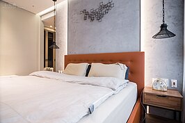 主臥室則花費較多心思裝潢。床頭部分沒有常看到的床頭櫃，而是高掛著四方角鐵所焊接成的廢鐵件感覺藝術品與鐵鍊吊燈，搭配油漆破刷方式所做成的牆面，讓這個空間溫馨中帶著冷冽的後現代風格。