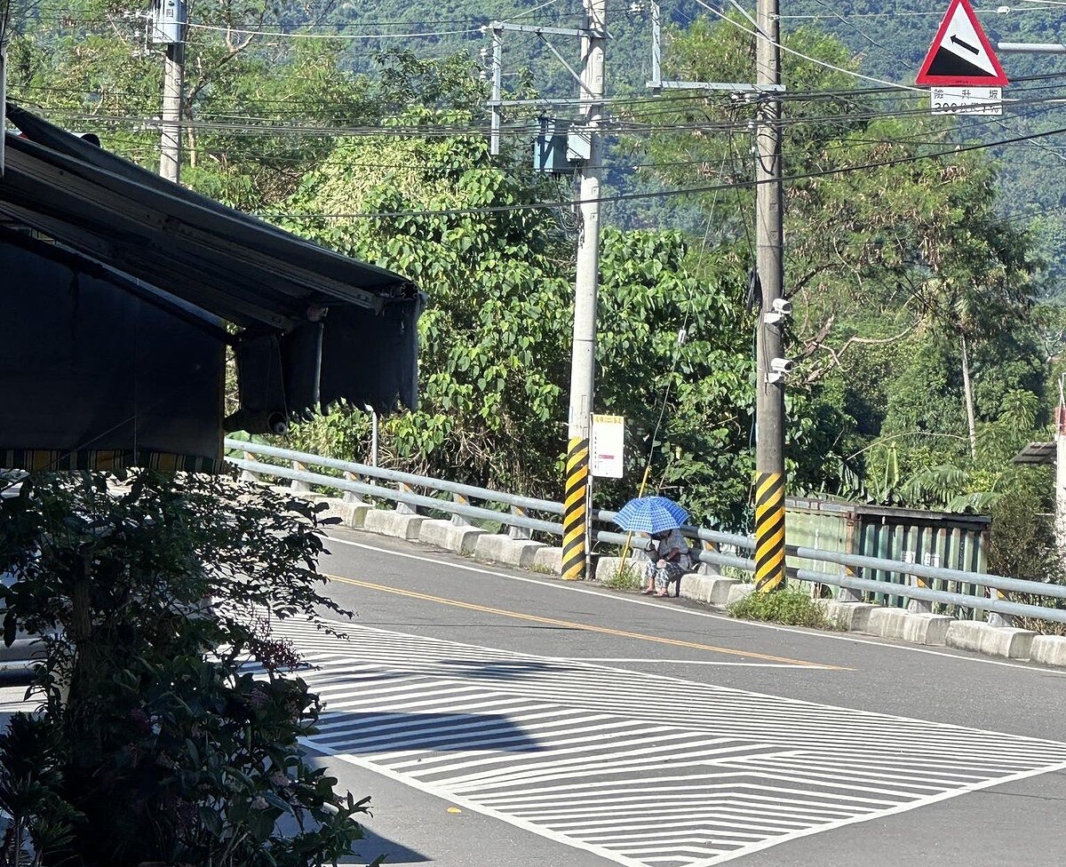 高雄六龜一名長者在烈日下撐傘坐在欄杆基座上等公車,畫面曝光後引起居民關切,盼市府重視偏鄉候車亭無法遮風避雨的問題。圖/記者徐白櫻翻攝